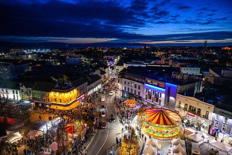 A lively city view of Galway at night, featuring illuminated streets, a carousel, and bustling crowds under a deep blue sky.