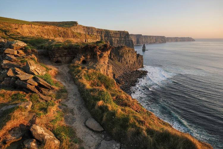 A dramatic coastal view of the Cliffs of Moher, with steep rock faces rising above crashing Atlantic waves under a bright sky.