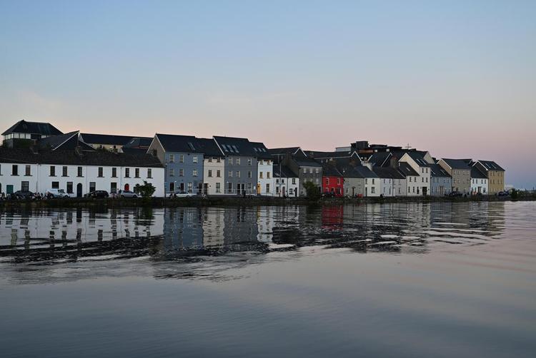 A tranquil view of the Long Walk in Galway, with colourful houses reflected in calm water under a soft evening sky.